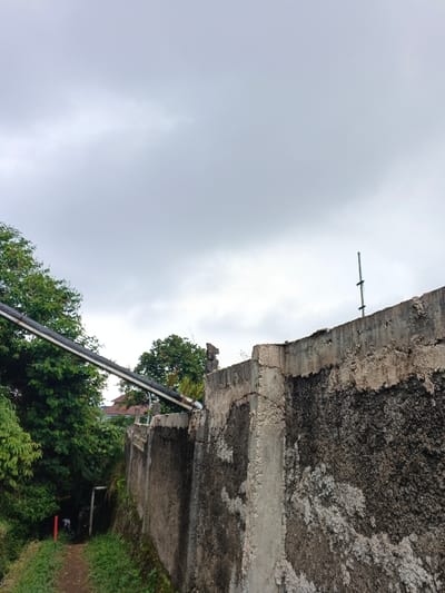This image depicts an outdoor scene in Koipa, Papua New Guinea, under an overcast sky. The upper two-thirds of the frame are dominated by a uniform light grey sky, indicating diffused lighting.
In the mid-ground, a weathered concrete retaining wall extends diagonally from the lower right to the mid-left. This wall exhibits significant deterioration, including spalling, visible cracks, and large patches of dark discoloration, suggestive of moisture or organic growth. Its top edge is irregular and broken in several places. A metal pole, light grey in color, is affixed vertically to the top of the wall on the right side.
To the left of the wall, a dark-colored pipeline, possibly conveying water or cables, runs diagonally downwards from the upper left, supported above a dense canopy of green trees and bushes. Below the pipeline and adjacent to the wall, a narrow, unpaved dirt path with reddish-brown earth is visible, flanked by green grass and vegetation.
Along this path in the distance, two individuals are present. The first, closer to the foreground, is wearing a light-colored shirt and dark trousers, facing away from the viewer. Further down the path, a second, less distinct individual is partially obscured by a dark, gate-like structure or frame. A vertical red marker or post stands beside the path on the left. Through the foliage in the background, a building with a reddish-brown roof can be partially seen.
