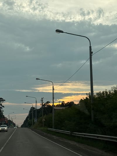 The image depicts a road scene in Timiryazevskoe, Russia, during what appears to be dusk or early morning. The sky is overcast with dramatic, textured clouds, tinged with the warm glow of the setting or rising sun peeking through a break in the clouds towards the horizon. A paved road dominates the foreground, with white lane markings visible. Several cars are present: a white car is in the left lane, and two other cars are further down the road, appearing as distant specks.
Along the right side of the road, a metal guardrail runs parallel to the asphalt. A steep embankment covered in dense green foliage rises behind the guardrail. Several tall, slender streetlights with curved arms are positioned along the road, casting no light as it is not yet dark enough. Electrical poles with wires are also visible, integrated with some of the streetlight structures. On the left side of the road, trees line the shoulder, and beyond them, structures and a sign can be partially seen. The overall impression is that of a rural or suburban road on the outskirts of a settlement, under a moody sky.