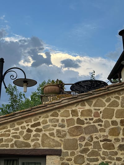 The image shows a stone wall with a terracotta tiled roof under a cloudy sky. On the roof, there is an old-fashioned lamp post with a spiral arm, a large terracotta pot with plants, and a black metal table with a plant growing from its center. To the right, a black chimney protrudes from a dark roof. The architecture suggests a rustic building, likely in a rural area. The image is taken outdoors, at eye level, and focuses on the upper portion of a building. The lighting suggests it is either early morning or late afternoon. The specific location in Cannara, Italy, is not explicitly visible, but the style of the building and stonework is consistent with rural Italian architecture.