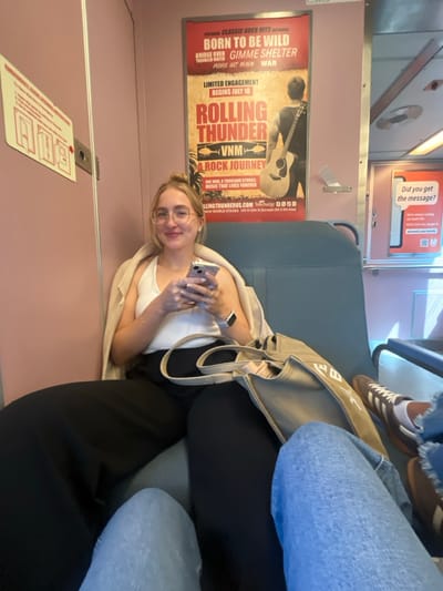 A young adult female with blonde hair, wearing glasses and a light-colored top with a beige jacket, smiles at the camera while seated on a commuter train. She holds a silver smartphone with both hands and has a smartwatch on her left wrist. Her legs are clad in black trousers, partially obscured by a light beige tote bag resting on the seat beside her. The train's interior features light pink-beige walls and blue-grey upholstered seats. Above the woman, a poster advertises "ROLLING THUNDER VNM: A ROCK JOURNEY" at "NEW WORLD STAGES 340 W 50th St" in New York City. To the right, a smaller sign visible states, "Did you get the message?" and includes "njtransit.com/myalerts," identifying the vehicle as an NJ Transit train. The lower legs of a second individual, wearing blue jeans and brown Adidas Samba sneakers, are visible in the foreground, suggesting they are the photographer. The scene takes place inside an NJ Transit train, consistent with transportation operating through Linden, New Jersey, United States.