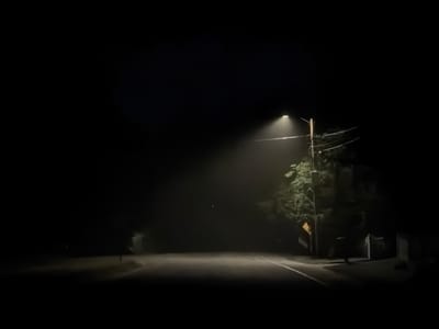 This image captures a dark, foggy night scene on a road in Hampton Bays, United States of America. A streetlamp on the right side of the road emits a bright, diffused light that illuminates the fog and a portion of the road. The road appears to be paved with white lines marking the lanes. To the right of the road, a utility pole with wires and a yellow sign are visible, along with some foliage from a tree. Further to the right, the indistinct outline of a building with a fence can be seen. The rest of the scene is enveloped in darkness, with only faint hints of distant objects. There are no people or vehicles clearly depicted in the image. The atmosphere is somber and somewhat mysterious due to the low visibility and the limited light.