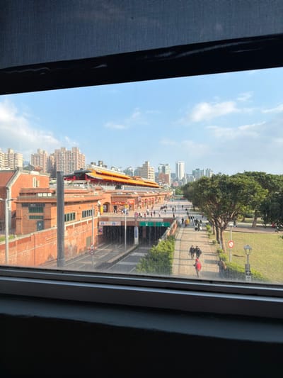 The scene viewed through a window shows a street with people walking and biking along the sidewalks. Brick buildings line the street, and in the distance, there are high-rise buildings. A temple-like structure is visible. Trees and greenery are present, contrasting with the urban environment. The sky is blue with scattered clouds.