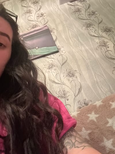 The image shows a close-up view of a person lying down. The person has dark hair and appears to be wearing a pink top. Part of their face, including an eyebrow and eye, is visible. A small part of an arm with a tattoo is also visible, along with a light brown rug patterned with stars. Behind the person are a wall with a floral pattern and a rectangular object which may be a mirror. There is no information to identify this as being Dobrovnitsa, Bulgaria.