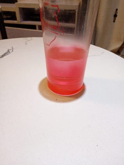 An indoor scene in Olambe, Nigeria, depicts a clear plastic cup containing approximately four ounces of translucent reddish-pink liquid, positioned centrally on a light-colored, textured horizontal surface, likely a table. Faint red branding is visible on the cup's upper side. A distinct shadow from the cup is cast to its right. A thin, dark, string-like object rests on the table surface to the left of the cup. The background is out of focus, revealing blurred angular structures consistent with shelving or furniture on the left, and a lighter, indistinct wall or large object on the right. No individuals, actions, or specific interactions are visible.