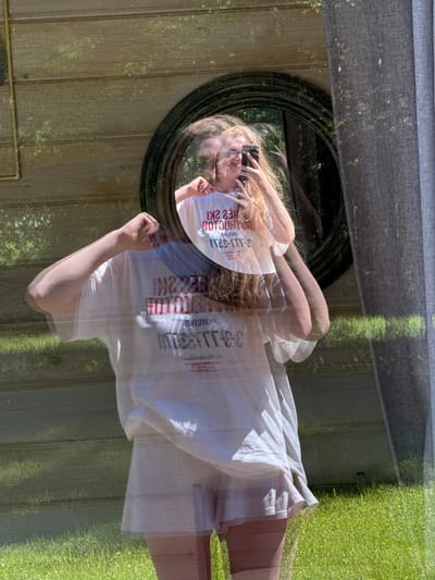A composite image depicts a mirror selfie captured through a reflective surface, presenting a female subject overlaid on an outdoor scene in Копнино, Russia. The subject, a female with long, light-colored hair, is positioned centrally, facing slightly left, taking a photograph of her reflection in a circular mirror with a dark, ornate frame. She holds a dark-colored smartphone to capture the image. She wears an oversized white t-shirt with red and black text, reversed in the mirror to read "SKI HOTLINE 3-111-S2H" and appearing as a translucent overlay on her form. Light-colored shorts are visible beneath the t-shirt, along with her legs, which appear to be covered by light-colored hosiery or leggings. The subject in the mirror reflection exhibits a smile. The background, visible through the subject's form and around the mirror, shows a brightly lit outdoor environment with a horizontally slatted wooden fence or wall on the left, vibrant green grass in the lower half, and a dark grey, pleated curtain on the right. Dappled sunlight suggests an overhead tree canopy, illuminating the wooden wall and grass.