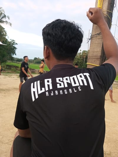 In Turumame, Indonesia, a young person is seated, viewed from behind. They are wearing a black t-shirt with white lettering that reads "HLR SPORT AJANGALE". Their right arm is raised, hand gripping a wooden pole to which a volleyball net is attached. In the background, other young people are visible in a sandy outdoor area, seemingly playing volleyball, with greenery and trees beyond. The sky is overcast.
