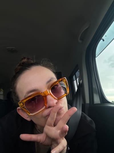 A photograph taken inside a vehicle, possibly in Kulaevo, Russia, although the location is not definitively identifiable from the image alone. A person with brown hair pulled back in a bun is seated, wearing orange-framed sunglasses. Their face is partially obscured by their hand, which is raised towards their mouth. The individual is wearing a black garment and is positioned in the driver's seat, evidenced by the seatbelt visible. The vehicle's sunroof is open, allowing a view of a cloudy sky. The interior of the car is a mix of gray and black materials. A passenger grab handle is attached to the roof near the top of the frame.