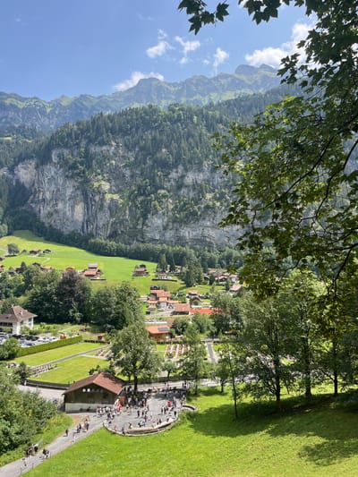 The image captures a scenic view of the Swiss Alps, likely from a high vantage point, overlooking a village nestled in a valley. The foreground is dominated by lush green grass and trees, with dappled sunlight casting shadows. In the middle ground, a cluster of traditional Swiss chalets with red-tiled roofs are spread across verdant fields, bordered by a sheer, imposing rock face covered in dense evergreen trees and a thin waterfall.
In the lower part of the image, a pathway winds through a grassy area, with several people walking and gathered. A small, wooden building with a red roof is situated near a group of people interacting. Further to the left, a parking lot filled with cars is adjacent to a manicured lawn and what appears to be a cemetery with rows of markers.
The background features a dramatic mountain range, with sharp peaks partially obscured by greenery and a clear blue sky with scattered white clouds. The overall impression is one of a serene, natural landscape combined with a well-maintained village, typical of the Bernese Oberland region.