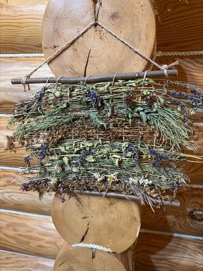 A bunch of dried herbs and flowers, including lavender, hangs from two horizontal wooden logs, which are part of a rustic wooden wall. The logs are secured by thick white rope. The herbs are tied to a thin branch, which is suspended by twine attached to the logs. The overall scene evokes a sense of natural, rural decoration.
