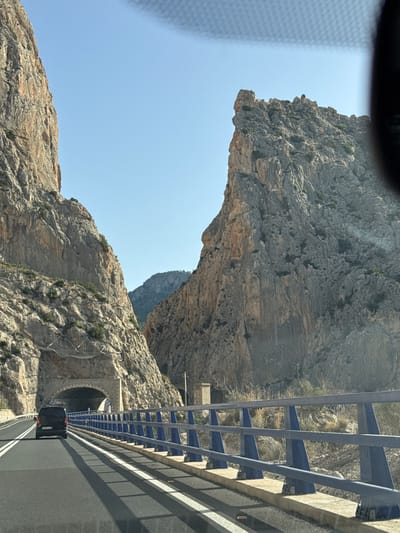 The image captures a view from a vehicle traveling on a road that passes through a dramatic mountain pass. On the left, a sheer rock face rises steeply, partially obscuring a tunnel entrance. On the right, another imposing rock formation with jagged peaks dominates the scene. A blue and grey metal guardrail runs along the side of the road, separating it from the mountain slope. A black van is seen ahead, driving towards the tunnel. The sky is a clear, light blue. The overall environment is mountainous and rugged. This location is known as the Bernia mountain pass, a scenic route near Altea, Spain, offering breathtaking views and challenging terrain.