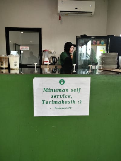 An indoor scene captures the counter area of a coffee shop or cafe, likely in Dramaga, Indonesia. A female barista, wearing a black cap and a green uniform, stands behind a dark counter. To her left, two plastic cups filled with layered beverages are visible. Behind her on the counter are a red blender and a black electric kettle. To her right, a glass-front refrigerator holds various items, and a tall stack of white plates is neatly arranged. An air conditioner is mounted on the light-colored wall above the counter.
In the foreground, a white sign with green text is taped to the dark green counter. The sign reads: "Minuman self service, Terimakasih :)" which translates to "Drinks self service, Thank you :)". Below this, it indicates "Bentokopi IPB", likely the name of the establishment. In the background, behind the counter, a "STAFF ONLY" sign is visible on a glass door, suggesting a restricted area. The reflections of people or objects can be seen in the glass surfaces, indicating a bustling or active environment. The overall setting appears clean and organized, typical of a service area in a commercial establishment.