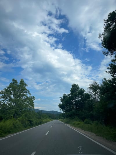 The image depicts a two-lane asphalt road stretching into the distance, flanked by dense green trees and vegetation. The sky is a mix of blue and white clouds. The scene appears to be a rural setting in Komarevo, Bulgaria, as suggested by the presence of rolling hills in the background. A portion of a car's hood with a Mercedes-Benz logo is visible in the foreground. There are no people or animals visible in the image. The overall impression is one of a quiet, scenic drive through the countryside.