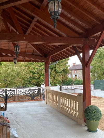 The image shows a covered outdoor patio area with a wooden roof and supporting beams. Two ornate lanterns hang from the ceiling. A decorative metal railing with a tree motif runs along the edge of the patio, behind which is a stone house with a tiled roof and blue shutters. To the right of the patio, there is a stone balustrade and a potted green shrub. A white sneaker and a wooden crate are visible on the concrete floor. The scene appears to be in Courpière, France, based on the architectural style of the house and the surrounding foliage.