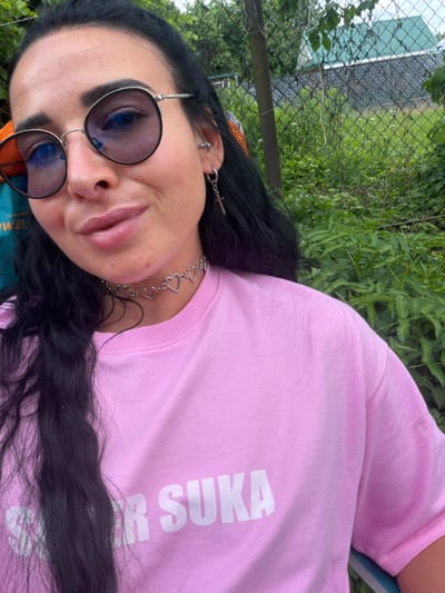 A medium shot photograph shows a woman with dark hair and sunglasses posing outdoors. She is wearing a pink sweatshirt with white lettering that appears to say "ER SUKA". She is also wearing a choker necklace made of small silver hearts and earrings. The background consists of a chain-link fence, lush green foliage, and a glimpse of a building with a green roof. The overall scene suggests a casual, outdoor setting. The location is identified as Bronnitsa, Russia.