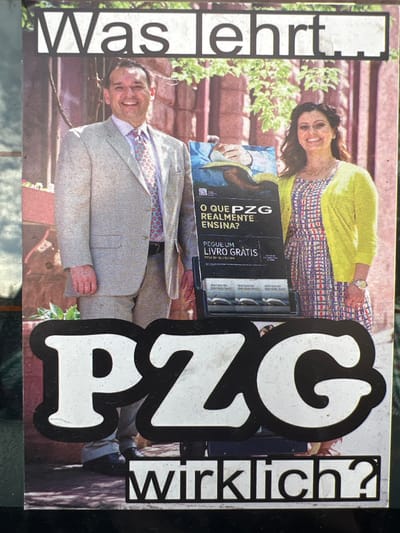 This image is a photograph of a printed poster located in Uccle-Ukkel, Belgium. The poster features two smiling Caucasian individuals standing behind a literature display cart. On the left, a man with short, dark hair wears a light-colored suit, a light shirt, and a patterned tie, smiling directly forward. To his right, a woman with long, dark, wavy hair wears a patterned dress and a bright yellow cardigan, also smiling directly forward. She wears a watch on her left wrist. Between them, a dark-colored literature display cart holds multiple booklets. A sign on the cart displays an image of hands holding an open book, along with Portuguese text: "O QUE PZG REALMENTE ENSINA?" (What does PZG really teach?) and "PEGUE UM LIVRO GRÁTIS" (Take a free book). The background within the poster depicts a reddish-brown brick wall partially covered by green foliage. Overlaid on the top of the poster is German text: "Was lehrt." Prominently displayed across the lower section are large white letters with a black outline, "PZG," followed by smaller black text on a white background, "wirklich?" The photograph itself shows a slight reflection on its left side and the texture of the printed poster.