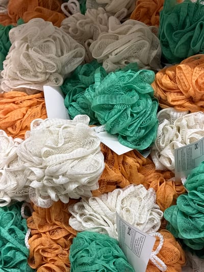 This image presents a close-up, overhead view of a retail display featuring a dense collection of mesh bath sponges, commonly known as loofahs, inside an IKEA store located in Corsico, Italy. The sponges are haphazardly arranged, appearing to be contained within a bin or basket. The color palette of the sponges includes white, orange-peach, and green. Several white product labels are partially visible embedded among the items. One visible label clearly displays the "IKEA" logo and product information, including "Made in China" and distribution details for "IKEA Ltd. 100 Avebury Boulevard, Milton Keynes, United Kingdom, MK9 1FH," alongside a copyright notice for "© Inter IKEA Systems B.V. 2015." No individuals, actions, or additional environmental details beyond the products are discernible within the frame.