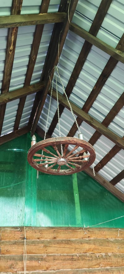 The image shows a rustic chandelier made from a wooden wagon wheel, suspended by chains from a wooden beam structure supporting a corrugated metal roof. Two light bulbs are visible on the chandelier. Behind the chandelier is a translucent green wall, and below that, a wall constructed of horizontal logs. The scene appears to be an outdoor or semi-outdoor covered area, possibly a porch or a barn. There are no people or animals visible in the image. The location is identified as Altayskoye, Russia.