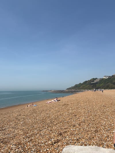 The image shows a sunny day at a pebble beach in the Folkestone and Hythe District, United Kingdom. The sky is predominantly blue. A body of water, likely the sea, runs along the length of the beach. The shoreline is covered in small pebbles and stones. Several people are on the beach, some sunbathing and others possibly walking. A green, overgrown hillside rises adjacent to the beach, and some buildings are visible near the top of the hill. A towel or fabric object rests on the pebbles close to the foreground.