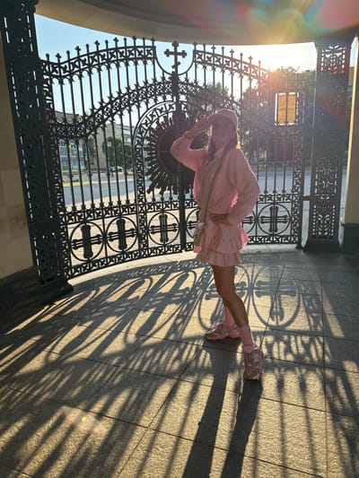 A woman in a pink outfit poses in front of a large, ornate black metal gate in Kirishi, Russia. The sun is setting, casting long shadows on the paved ground. The gate features intricate scrollwork and circular designs with crosses. In the background, a street with traffic lights and buildings is visible through the gate. The woman is wearing a pink collared shirt, a ruffled skirt, pink socks, and pink sneakers, along with a pink baseball cap. She has one hand on her hip and the other raised to her forehead, as if shielding her eyes from the sun. A small, quilted handbag hangs from her shoulder.