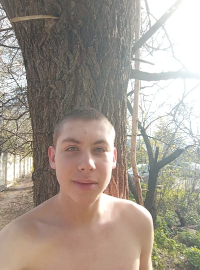 This image captures a young man outdoors in what appears to be a natural or semi-natural setting, possibly in совхоза "Воронежский", Russia. The photo is oriented ninety degrees clockwise. When viewed upright, a young man, shirtless with short light-brown hair and striking light-green eyes, is prominently featured on the left side of the frame. He looks directly at the camera with a calm, subtle smile, his face illuminated by bright sunlight which causes some glare on his skin.
To his right, a large tree trunk with rough, textured bark dominates the middle and right side of the composition. The man appears to be standing very close to or leaning against this tree. In the background, a bright, clear sky is visible through a dense network of bare tree branches, suggesting a time of year when deciduous trees are without leaves, possibly early spring or late autumn. Some faint green foliage can be seen in the lower background, along with a glimpse of an indistinct fence or structure in the upper left background. The strong sunlight suggests it is daytime, casting bright highlights across the scene. There is no visible text.