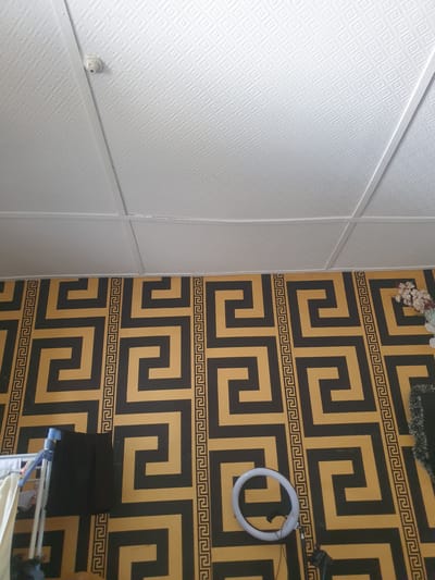 The image depicts an interior scene, likely a room, with two distinct wall coverings. On the left, the wall is covered in white, textured panels with a repeating diamond pattern. A small white smoke detector is visible on this wall. On the right, the wall features a bold geometric wallpaper with a pattern of interlocking black and gold squares and Greek key motifs.

In the upper right corner of the wallpapered wall, a small cluster of artificial flowers can be seen. Hanging on the wall is a white circular object, possibly a light ring or a towel holder. Below this, a dark object, perhaps a piece of clothing, is draped. In the lower right of the frame, part of a clothes drying rack is visible, with some white fabric draped over it.

The lighting appears to be artificial and diffused, suggesting it is either daytime indoors or nighttime. There is no indication of weather or time of day beyond the indoor setting. The scene is static, with no people or obvious activity taking place, creating a sense of stillness. The location is identified as Fajo 2 Division Range, Nigeria.