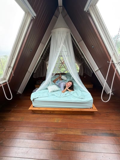 A woman lies in a bed draped with a white mosquito net in a room with wooden walls and floors. The bed is covered in light blue bedding and several pillows. The room features slanted ceilings with windows offering a view of greenery outside. White beaded cords hang near the windows. The woman is looking up towards the camera.