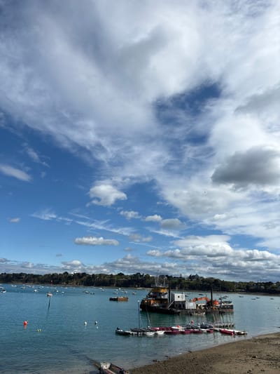 The image shows a view of the port of Dinard, France, located at coordinates (latitude: 48.634, longitude: -2.05). The scene is dominated by a bright blue sky with white cumulus clouds scattered across the expanse. The water in the port is a tranquil turquoise, with several boats and yachts anchored in the bay.  A large, yellow-and-white dredging vessel sits in the center of the image, with several smaller boats surrounding it. The shore in the foreground is a sandy beach with a rocky edge, and the background shows a lush, green hillside with trees and buildings. The image was taken during the day, and the sunlight creates a warm, inviting atmosphere.