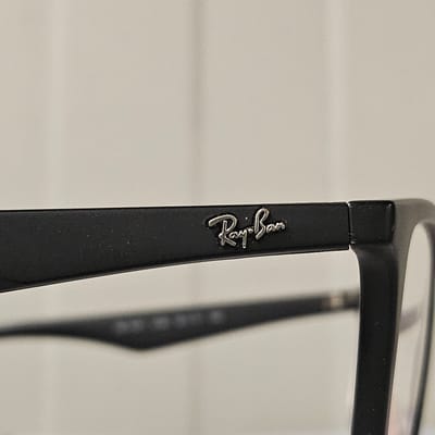 A close-up, eye-level, medium shot shows a matte black eyeglass frame with the silver Ray-Ban logo embossed on the exterior temple. The logo is in the center of the frame's temple and is slightly angled toward the viewer. The background is a plain, light-colored surface, possibly a wall. The image does not provide any information about the location in the city of Nowa Wieś Książęca, Poland.
