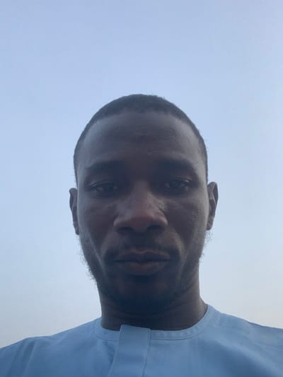 The image depicts a close-up portrait of a Black man, taken in a selfie style. He is looking directly at the camera with a neutral expression. He has short black hair and a short beard. He is wearing a light blue, long-sleeved shirt. The background is a clear blue sky. The coordinates provided place the image in Sokoto, Nigeria, likely taken outdoors.