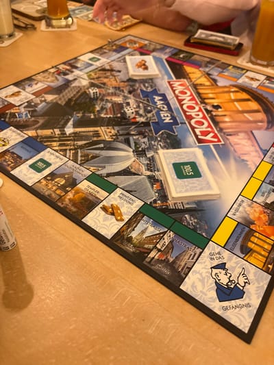 A customized "MONOPOLY AACHEN" board game is laid out on a light-colored wooden table. The game board features various Aachen landmarks and street names such as "Rathaus," "Aachen Hauptbahnhof," "Templer-Graben," "Pontstrasse," "Annastrasse," and "Elisenbrunnen," along with designated spaces like "Gefängnis" (Jail) and "Printen-Meldung." Two white dice are visible on the board, displaying faces indicating a roll in progress. A metallic game token rests on a property space. Two stacks of game cards are present; one stack prominently displays a green "100.5" logo on its top card. Two brown, almond-shaped cookies, identified as Aachener Printen, lie on the "PRINTEN-MELDUNG" space. In the background, partially visible human hands suggest active players. Two beverages are on the table: a clear glass with a light-colored liquid on the left and an amber-colored liquid in a glass on the right, both on coasters. A stack of currency, possibly US dollars, is visible beneath a dark-colored smartphone to the upper right. A white cylindrical object is partially visible on the far left edge of the table.