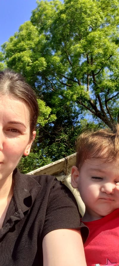 The media file is a low-angle, close-up photograph featuring two individuals outdoors, likely a mother and her child. The dominant elements are the lush green foliage of a tree overhead and the bright blue sky. In the foreground, a person's face is visible, with dark hair and brown eyes looking towards the camera. To the right, partially obscuring the background, is another person, a child, with light brown hair and closed eyes. The child is wearing a red shirt with a star graphic. Both individuals appear to be lying down, with the camera angled upwards from a low vantage point. The overall lighting suggests a sunny day, possibly in the late afternoon or early morning due to the warm tones. The setting appears to be a natural outdoor environment, with the abundance of green leaves indicating a warm season. The relaxed posture of the subjects and the casual nature of the shot suggest a moment of leisure and connection. There is no visible text in the image.