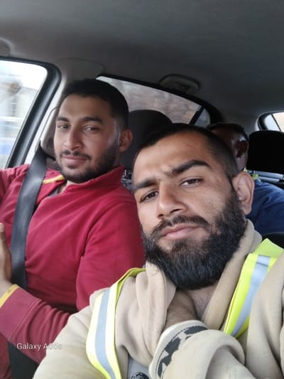 The scene depicts three adult males within the interior of a vehicle, located in Dobrevtsi, Bulgaria. In the foreground, on the right, a male subject with dark hair and a prominent dark beard and mustache wears a beige hooded sweatshirt overlaid with a yellow and grey reflective safety vest; his gaze is directed towards the camera. To his immediate left, a second male with dark hair and a trimmed beard is seated, wearing a red long-sleeved garment with a visible black seatbelt diagonally across his chest and left shoulder; he also looks towards the camera. A third male is partially visible in the vehicle's background, showing the back of his head and a portion of a blue shirt. The vehicle's interior components, including window frames and the headliner, are visible. The exterior environment seen through the windows is blurred. A "Galaxy A05s" watermark is present in the lower-left corner of the image.
