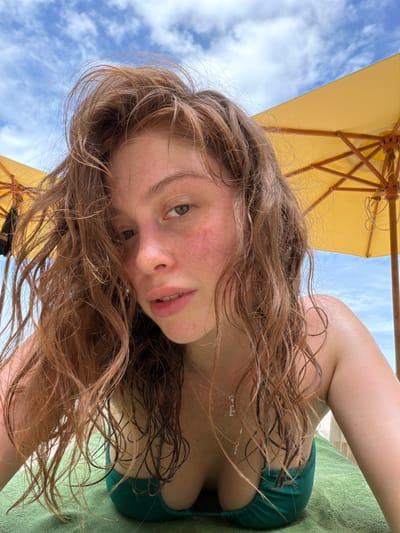 A young woman with auburn hair and freckles lies on a green towel under a yellow umbrella on a beach in Ao Cho, Thailand. The sky above is blue with scattered clouds. She is wearing a dark green bikini top and a silver necklace. Her hair is wet and windswept. The image is a close-up selfie, taken from a low angle, focusing on her face and upper body. The sun is bright, casting a warm glow on her skin.