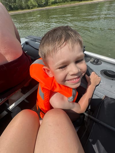 The media file is a portrait-oriented, close-up, outdoor, candid photograph of a young boy in an orange life jacket, smiling at the camera. He is likely on a boat, as indicated by the presence of the boat's interior structure, water, and a railing. The boy's face is wet, suggesting he may have been swimming or splashed. He has light brown hair and is wearing a dark red top. To the right of the boy, a person's legs, presumably an adult, are visible, also wet. The background shows a body of water with a green, treed shoreline. The lighting suggests it is daytime, likely sunny, with the sun casting shadows. The scene evokes a sense of fun and enjoyment, typical of a day spent on the water. No visible text is present in the image.
