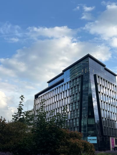 The image shows a modern glass-fronted office building against a cloudy blue sky. The building has a dark facade with many windows. In the foreground, there are trees and foliage. A blue sign with "GHELAMCO" written in white letters is visible on the lower part of the building. The overall scene suggests an urban environment, consistent with the location of Saint-Josse-ten-Noode - Sint-Joost-ten-Node, Belgium, known for its mix of residential and business properties. No people are visible in the image.