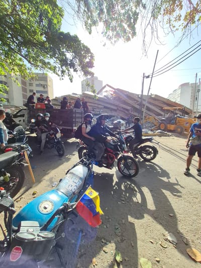 The image captures a scene of destruction and recovery in Caraballeda, Venezuela, following a natural disaster. In the background, a collapsed building, with its concrete beams and debris scattered across the ground, dominates the view. A yellow backhoe loader, with the "DEERE" logo visible, is positioned near the wreckage, suggesting ongoing cleanup or rescue efforts. Several people are present, some wearing vests and appearing to be part of a rescue or recovery team, while others are observing the scene.

In the foreground, motorcycles are parked on a paved surface, some with riders wearing helmets, indicating a mode of transportation for those present. A blue motorcycle in the foreground features a Venezuelan flag attached to its rear, a symbol of national pride amidst the difficult circumstances. The presence of power lines and a street lamp suggests an urban setting. The overall atmosphere is one of devastation, with the clear sunlight highlighting the debris and the ongoing efforts to address the aftermath of the disaster.