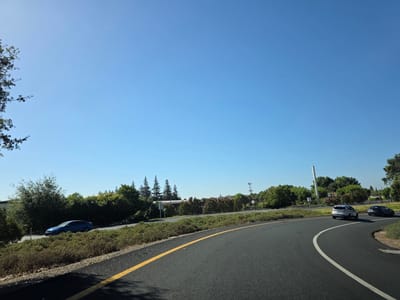 In Elk Grove, California, on a clear day with a vibrant blue sky, a road curves to the right, bordered by low green bushes and trees. Two cars are visible on the road: a silver SUV and a darker car following behind. To the left of the road, a bright blue car is parked near a line of trees. In the distance, a cell tower stands tall amidst the greenery. The image captures a typical highway scene in a suburban area.