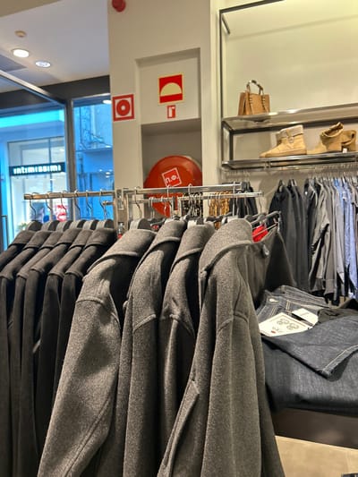 The image, taken inside a clothing store in Chalkida, Greece, captures a retail environment filled with garments and safety equipment. In the foreground, a rack holds numerous dark grey or charcoal long-sleeved garments, likely coats or thick sweaters, stacked neatly. Behind this rack, other clothing items, including dark shirts and trousers, are displayed on hangers. To the right, folded denim garments with "ZARA" tags and other symbols are visible.
On the wall in the mid-ground, a red fire hose reel with the Greek text "Π.Φ." (P.F.) is prominently displayed. Above it, a red and white sign indicates a fire extinguisher. Another red and white circular sign is below, possibly related to fire safety.
Further back and to the left, a display shelf showcases a pair of light brown ankle boots and a matching handbag. The background features reflective surfaces, possibly large mirrors or shop windows, which show reflections of interior lighting and another shop sign. Reflected on what appears to be a shop window or a polished floor, the word "intimissimi" is visible, indicating a nearby store.
No people are visible in the image. The scene is brightly lit by artificial light, suggesting an indoor setting during operational hours. The overall impression is that of an organized retail space, with a focus on clothing displays and adherence to safety regulations.