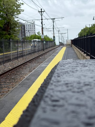 The image depicts a train station in Boston, Massachusetts, with the view along the tracks. The platform is wet, indicating recent rain. A yellow safety line marks the edge of the platform. Electrical wires and support structures are visible overhead, characteristic of a train line. In the background, buildings and trees are visible. The photo is taken at an angle from near the front of the platform which gives a strong depth of field.