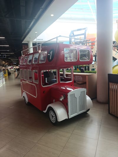 The image shows a red double-decker bus replica, likely a children's play structure, located indoors in a brightly lit mall. The bus is the primary subject, with its iconic design, including multiple windows on both levels and a large number "1" on its side. The setting appears to be a shopping center with a tiled floor, a high ceiling with exposed beams and skylights, and some merchandise displays visible in the background.
To the right of the bus, a portion of a playground structure is visible, with colorful elements and a large cylindrical pillar. Behind the bus, there are tables and chairs, suggesting a seating area within the mall. The lighting indicates it is daytime, with natural light filtering through the skylights. There is no indication of specific weather conditions.
The bus itself is red with white accents on the fenders and grille. The windows are clear, and some seats are visible inside. The overall impression is a cheerful and family-friendly environment. No people are clearly visible interacting with the bus, but the presence of a seating area and playground suggests it's a place for families.
The only visible text is on a sign in the background, which appears to be in a foreign language, possibly related to store names or directional information within the mall. The location context is Naro-Fominsk, Russia.