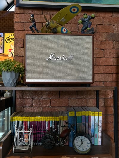 This image shows a close-up, top-down view of a collection of decorative items and books on a brick wall shelf. The primary subject is a vintage-style Marshall speaker, positioned in the center, with its iconic grille pattern and logo prominently displayed. To the left of the speaker, there are two small figurines: one of a superhero (likely Hulk) in a dynamic pose and another of Captain America. There is also a model airplane with a yellow fuselage and a circular red and blue emblem.

The right side of the image is dominated by a stack of books, many with yellow spines labeled with numbers, suggesting a series or catalog. Some of the book titles are visible, including "MOBILE SUIT GUNDAM UC" and other Thai titles. Interspersed with the books is a decorative metal clock with Roman numerals. A small metal sculpture of a bicycle or motorcycle is also present near the books. At the bottom, a gray geometric pot holds a small green plant with red-tinged leaves. A partial view of a yellow sign with "CAF Groo" is visible at the very bottom left. The brick wall provides a rustic background for all the items.

The scene appears to be indoors, possibly in a cafe or a home setting, given the mix of decorative and functional items like the speaker and books. The lighting suggests it could be daytime, but no specific weather cues are evident. The arrangement evokes a sense of personal collection and display, with a mix of pop culture and literary elements.