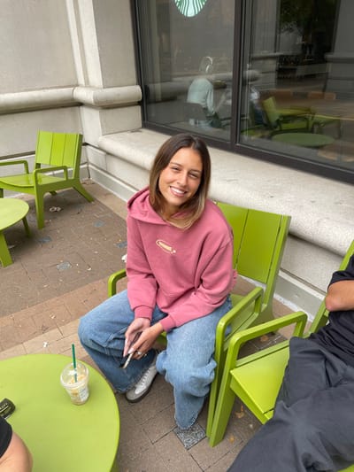 An outdoor scene depicts a young adult female seated in a lime green metal chair on a paved patio. She is smiling directly at the camera, wearing a dusty rose hooded sweatshirt with a "BROWNIE" logo and light blue denim jeans. Her feet are clad in light-colored sneakers, and she holds a gold/silver-colored smartphone with both hands. To her left, an iced coffee in a clear plastic cup with a green straw rests on a circular lime green metal table. To her right, a portion of another individual, wearing a black short-sleeved shirt and dark pants, is visible, also seated in a similar lime green chair. In the background, a building facade with light-colored stone or concrete cladding features large plate glass windows. The green siren logo of Starbucks is visible through the window, indicating the establishment. Reflections in the glass show interior details, including a person wearing headphones seated inside. The ground is paved with brown and grey interlocking pavers, with scattered organic debris. Several other unoccupied lime green chairs and a table are present in the immediate vicinity. This scene is located on an outdoor patio adjacent to a Starbucks establishment in an urban environment within Arlington, United States.