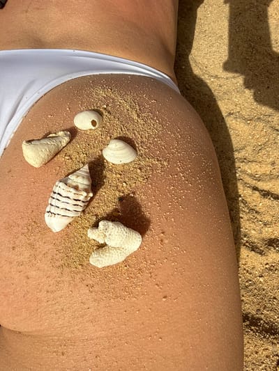 An overhead, close-up shot captures a person lying on a sandy beach in Safaga, Egypt, likely sunbathing or relaxing. The primary focus is on the upper part of a human leg and hip, which is partially covered by a white swimsuit bottom. The skin is tanned and has specks of sand clinging to it, indicating direct contact with the beach.

Arranged on the person's skin are several seashells and a piece of white coral. From left to right, a distinct piece of white, branching coral is visible. Next to it, a large, ornate conch shell with black striped markings rests. Further right and slightly above, there's a textured, light-colored shell. Below it, two smaller, smoother white shells are visible, one of which has a small, circular hole. Grains of sand are scattered across the skin, particularly around the shells and coral.

The lighting is bright and sunny, casting distinct shadows on the sand beneath the person, suggesting a clear day. The sand itself is golden-brown and coarse-grained. No text is visible in the image.