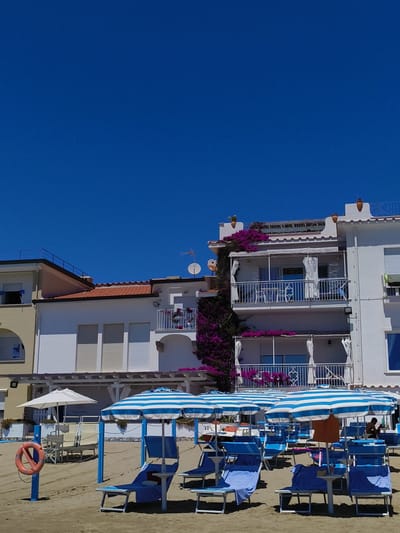 The image depicts a section of beach in Sperlonga, Italy, characterized by a clear blue sky dominating the upper portion. In the mid-ground, several buildings with white facades and balconies are visible, adorned with vibrant purple bougainvillea. These buildings appear to be hotels or apartments, with multiple stories and visible windows and shutters. Some balconies feature white curtains and outdoor furniture.

In the foreground, a beach scene is laid out. Numerous beach chairs and umbrellas, many with blue and white stripes, are arranged on the sandy shore. A few blue lounge chairs are occupied by people, though their faces are not clearly discernible. One person is seated in a chair, partially obscured by an umbrella, and another figure is visible further back. A life preserver ring, colored orange, is attached to a blue pole on the left side of the image. Beach umbrellas and chairs are set up in neat rows, suggesting a commercial beach operation. The sand is a light brown color. The overall impression is one of a sunny, relaxed day at the seaside.