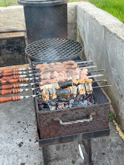 The image shows a barbecue grill in Новая Чернушка, Russia. Skewers of meat and fish are being cooked over hot coals. The grill is made of metal and has a handle on the side. The background is a concrete surface.