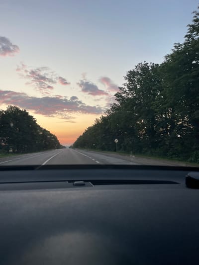 This media file depicts a view from inside a vehicle looking out at a road lined with trees on either side. The scene is set during what appears to be sunset or sunrise, as indicated by the warm hues of orange and pink in the clouds against a pale blue sky. The road itself is paved and has white lane markings.
In the foreground on the right side of the image, a portion of the vehicle's interior is visible, specifically a dark-colored dashboard or pillar. On the left side, a dense canopy of green trees flanks the road. The sky above the trees shows scattered clouds tinged with the colors of the setting or rising sun.
A single, round street lamp on a pole is visible in the distance along the side of the road, set against the backdrop of the trees and sky. The overall mood of the image is serene and contemplative, perhaps capturing a moment of travel or reflection. There is no visible text in the media file. The location context provided is Cricova, Moldova.