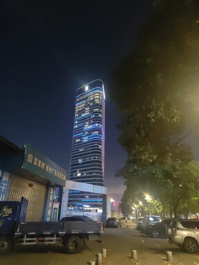 The image captures a nighttime scene on a street in Nanhai District, China. A tall, modern skyscraper with blue LED lighting outlining its floors dominates the left side of the frame, topped with a glowing white "H" logo. In the foreground, a blue flatbed truck is parked on the right side of the street, its cargo bed empty. To the left of the truck is a building with a closed roll-up door and a sign above it bearing Chinese characters, including "蓝果树" (Lan Guo Shu) and "板材广东运营总部" (Ban Cai Guang Dong Yun Ying Zong Bu), which translates to "Lan Guo Shu Wood Panels Guangdong Operations Headquarters." Across the street, several parked cars are visible, including a silver SUV in the right foreground. The street is lined with trees, illuminated by streetlights, casting a warm glow on the asphalt. The sky is dark blue, suggesting it is late evening or early night. A single person is seen walking down the sidewalk in the distance.