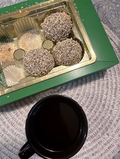 A top-down shot shows a green box of chocolates next to a black mug of coffee. The box contains an assortment of round, coconut-covered chocolates. Three of the chocolates are dark and covered in finely shredded coconut, while others are lighter and appear to be filled with white coconut. The chocolates are arranged in a plastic insert within the green box, which is open to reveal its contents. The mug of coffee is full and black, casting a slight reflection on its surface. The items are placed on a woven placemat with a circular pattern, which is on top of a patterned tablecloth. The lighting suggests an indoor setting, possibly a home or cafe. The scene evokes a sense of a relaxed moment, perhaps a morning or afternoon break. No people are visible in the frame. There is no discernible text in the image.
