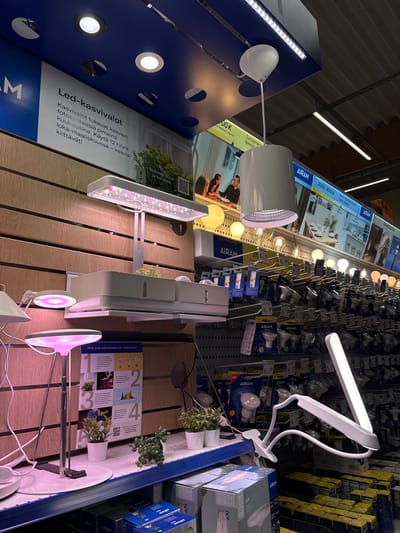 The image shows a retail display of various types of lighting, with a focus on plant lights. In the foreground, several modern plant lamps are showcased. One is a slim, white LED lamp with a flexible arm and clip, designed for attaching to shelves or desks. Another features a wider, rectangular LED panel emitting a pinkish-purple light, typical for plant growth. Further back, a circular pink light is also visible, as well as other smaller white LED downlights integrated into a blue display panel.
The background reveals shelves stocked with numerous light bulbs, primarily from the brand AIRAM. Boxes of various light bulbs are stacked high, indicating a well-stocked retail environment, likely an electronics or home improvement store. Some of the packaging displays information in Finnish, such as "AIRAM," "EASY," and "LIHTNE."
A prominent sign in Finnish, with the heading "Led-kasvivalot," translates to "LED Plant Lights." It explains that these lights support plant photosynthesis during dark months and recommends using them for 12 hours a day from October to March. Another sign offers a QR code to "Skannaa koodi ja lue lisää!" which means "Scan the code and read more!". A display behind the plant lights shows illustrated guides with numbers 1, 2, 3, and 4, possibly explaining different aspects of plant lighting or types of bulbs.
While no people are clearly visible in the image, the setting suggests a commercial space. The lighting appears to be artificial, typical of an indoor store. The overall impression is one of a well-organized display designed to inform and attract customers interested in plant cultivation or specialized lighting solutions.