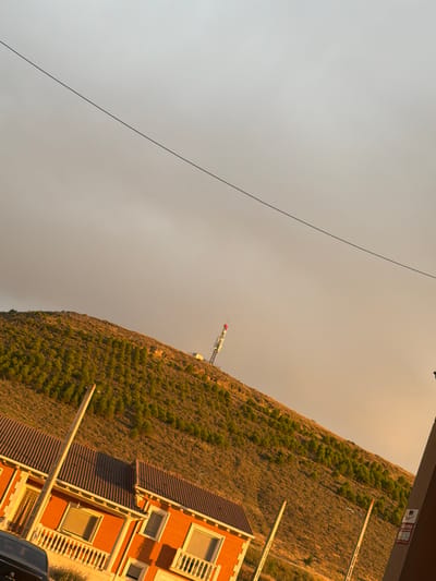 The image, taken in Vallelado, Spain, captures a hillside covered in dense, green trees, receding into the distance under a hazy, golden sky. The hillside is intersected by a series of wooden poles, some of which are visible in the foreground. A communication tower with red and white sections stands prominently on the crest of the hill. In the lower left of the frame, a portion of an orange brick building with a tiled roof is visible. The building features several windows with white railings and a balcony. A black car is partially visible in the bottom left corner. The overall scene is characterized by warm, diffused lighting, suggesting it was taken during sunset or sunrise. A thin, dark line, possibly a power line or cable, diagonally bisects the sky.